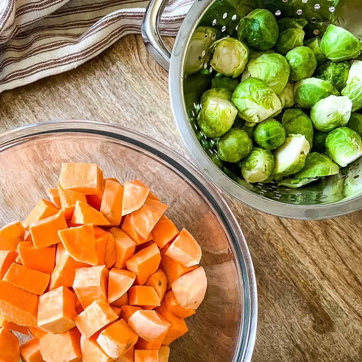 One glass bowl filled with cubed sweet potatoes and a colander with freshly washed brussels sprouts